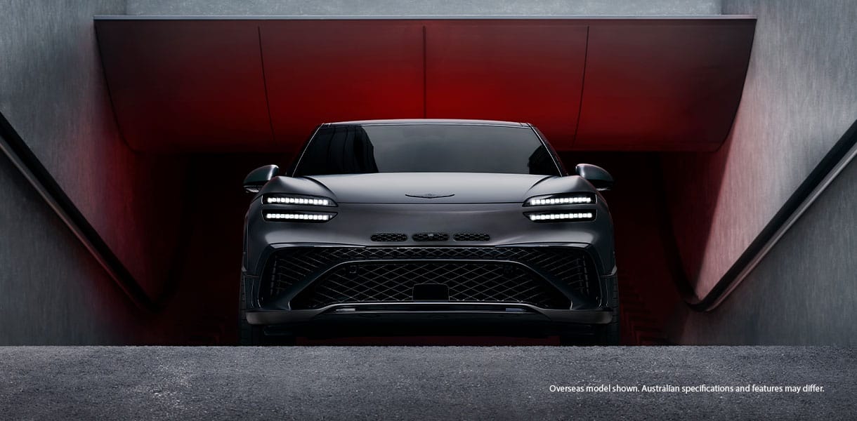 Front view of a Genesis GV60 Magma concept electric SUV emerging from a sleek concrete garage, illuminated by dramatic red ambient lighting, with distinctive LED headlights and a bold grille design. 