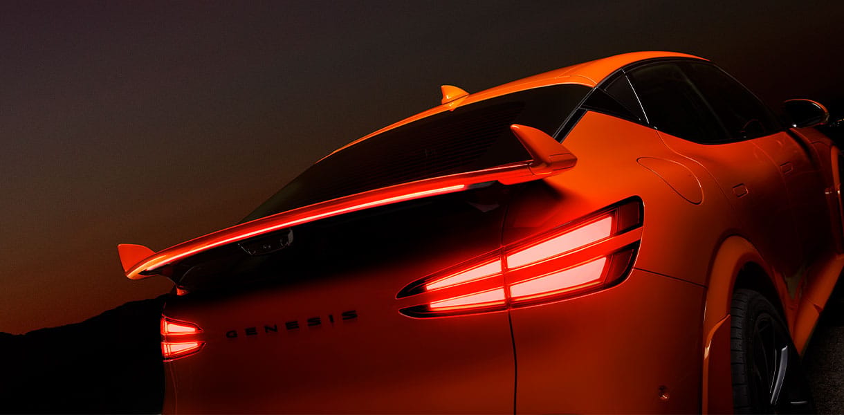 Close-up rear view of an orange Genesis GV60 Magma highlighting the illuminated dual-line LED tail lights and integrated rear spoiler, set against a dark background with dramatic lighting that emphasises the vehicle’s sculpted design and bold contours.