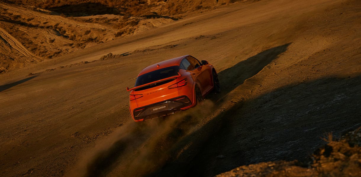 Orange GENESIS GV60 Magma SUV driving up a sandy desert hillside at sunset, kicking up dust with warm golden light casting long shadows across the rugged terrain.