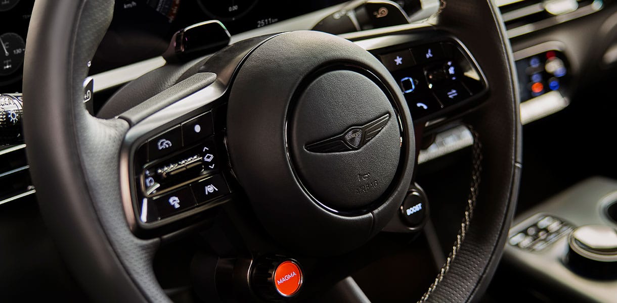 Close-up of a GENESIS steering wheel featuring paddle shifters and controls, highlighting the virtual gear shift interface with a prominent red “MAGMA” drive mode button and modern illuminated dashboard displays in the background.