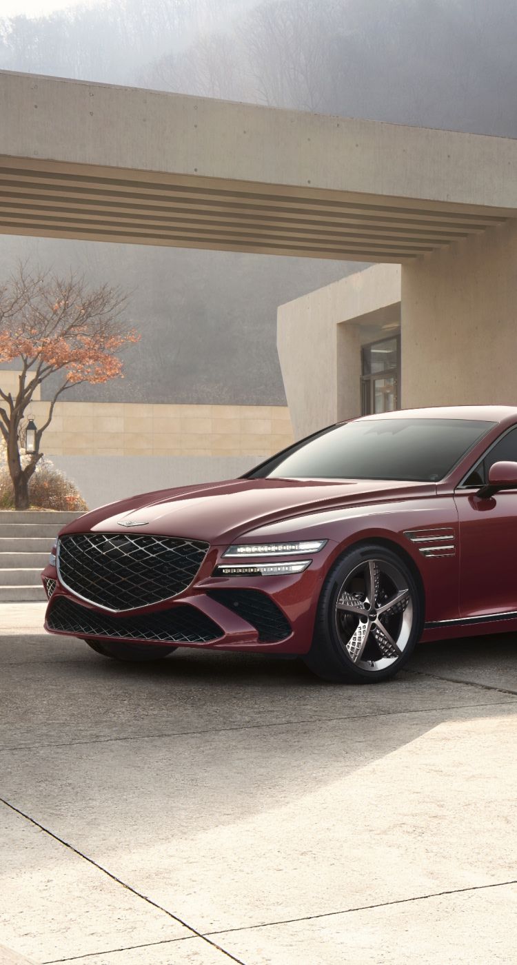 Genesis G80 luxury sedan in a striking red finish, parked against a modern architectural backdrop with sleek concrete structures and landscaped greenery.