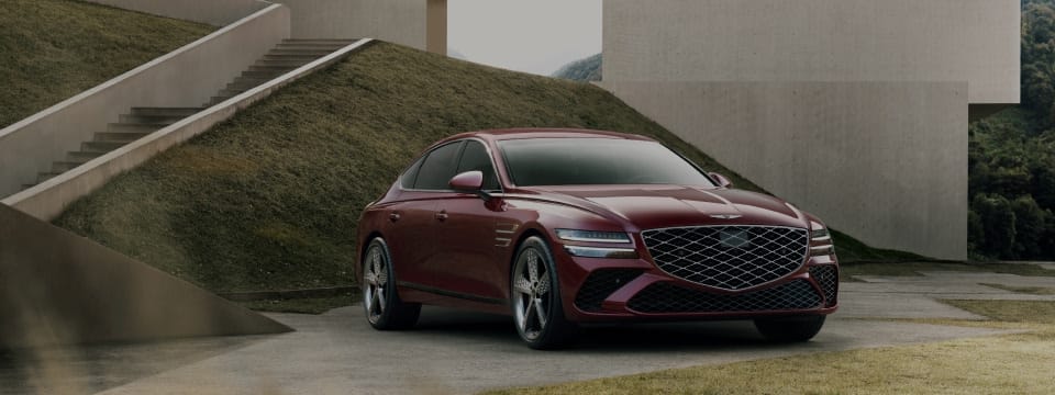 Genesis G80 luxury sedan in a striking red finish, parked against a modern architectural backdrop with sleek concrete structures and landscaped greenery.