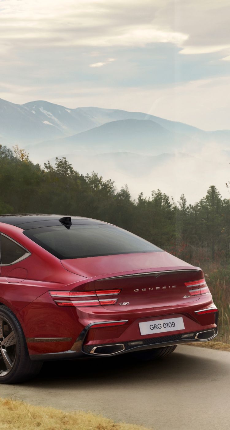 Genesis G80 luxury sedan in a bold red finish, driving along a road with a scenic green landscape and mountains in the background.