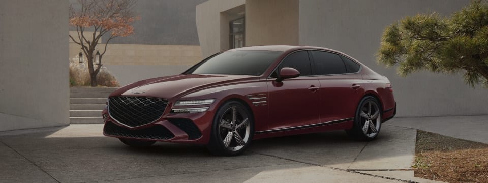 Genesis G80 luxury sedan in a striking red finish, parked against a modern architectural backdrop with sleek concrete structures and landscaped greenery.