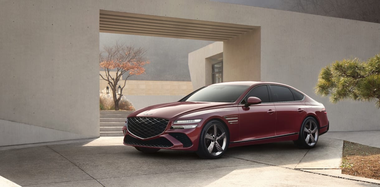 Genesis G80 luxury sedan in a striking red finish, parked against a modern architectural backdrop with sleek concrete structures and landscaped greenery.