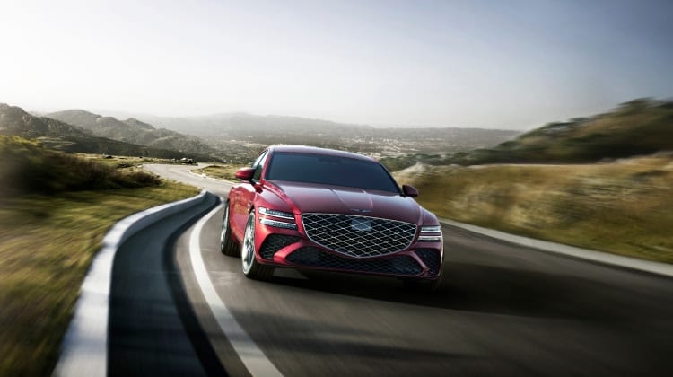 Genesis G80 luxury sedan in a bold red finish, driving along a road with a scenic green landscape and mountains in the background.