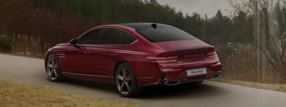 Genesis G80 luxury sedan in a bold red finish, driving along a road with a scenic green landscape.
