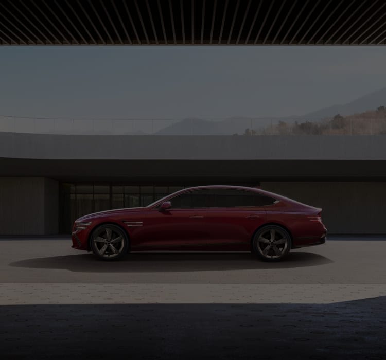 Genesis G80 luxury sedan in a striking red finish, parked against a modern architectural backdrop with sleek concrete structures and landscaped greenery.