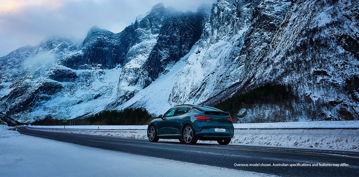 Genesis GV60 electric SUV driving on a snow-covered mountain road, with steep, rugged cliffs and pine trees in the background under a cold, overcast sky. Snow blankets the landscape, and the car is shown from the rear three-quarter angle. Small text in th