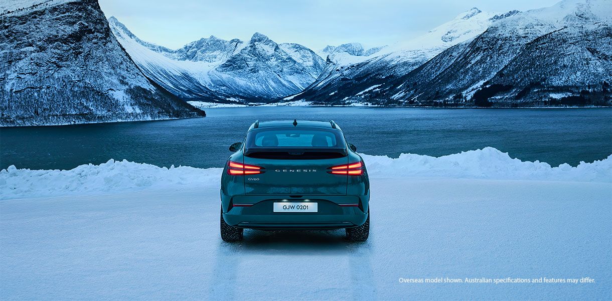 Genesis GV60 electric SUV viewed from the rear on a frozen lake, surrounded by snowy mountains and a scenic winter landscape.