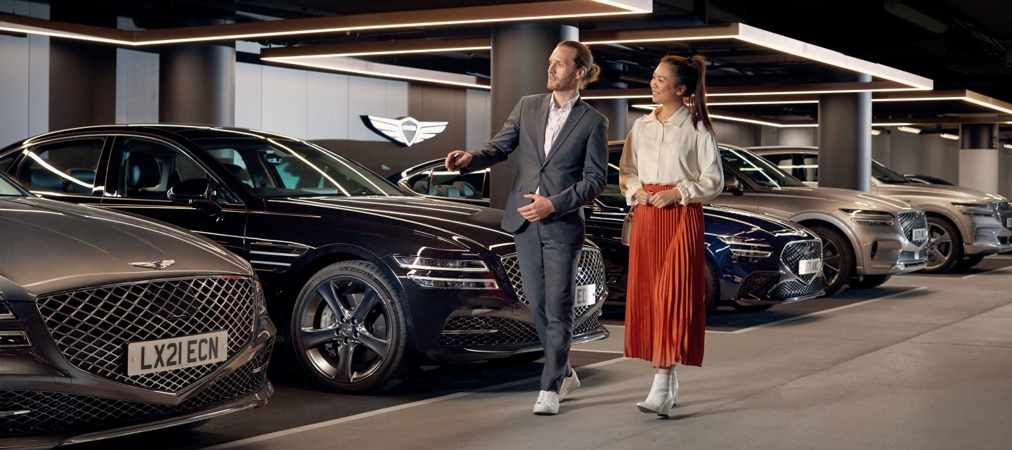 A friendly, smartly-dressed Genesis Assistant stands next to a gleaming car in a studio talking to a customer