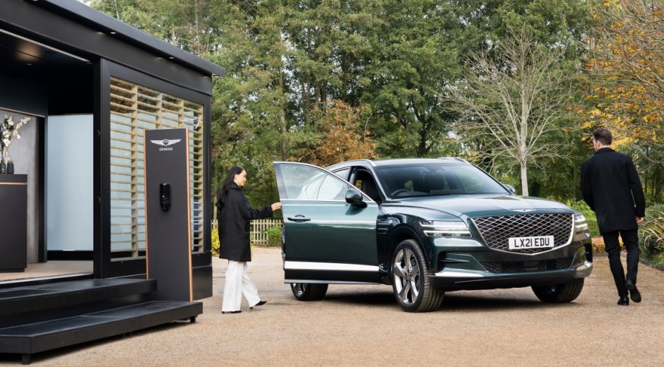Outside a Genesis Mobile Lounge a woman  opens the door of a large dark green SUV. A man walks around the other side.