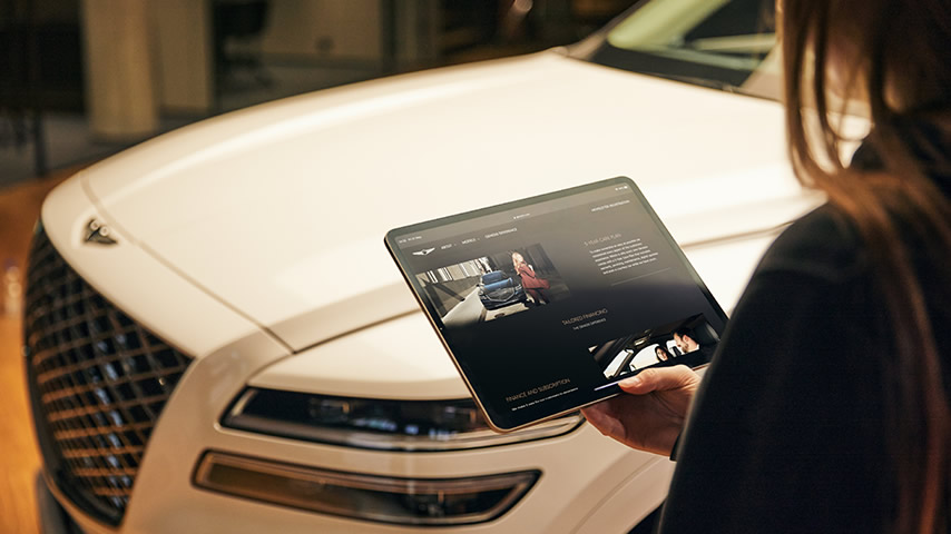 In front of a white Genesis a friendy woman holds an iPad with finance options on the screen