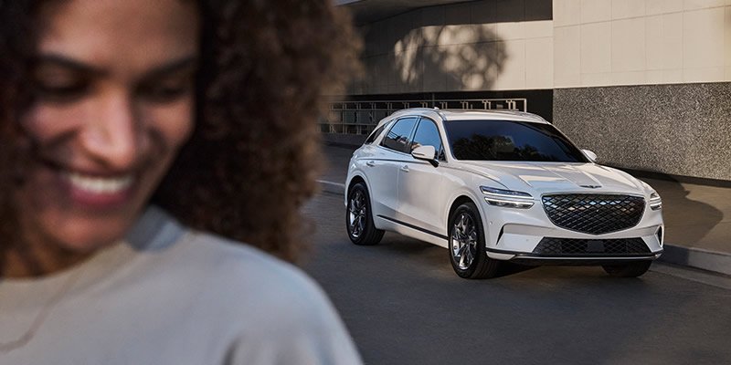 In evening sunlight a casually dressed woman smiles in the foreground with a new electric SUV behind her shoulder
