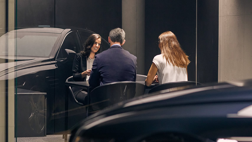 A Genesis Personal Assistant smiles at a smart couple in an office. A beautifully-lit saloon is reflected in the window 