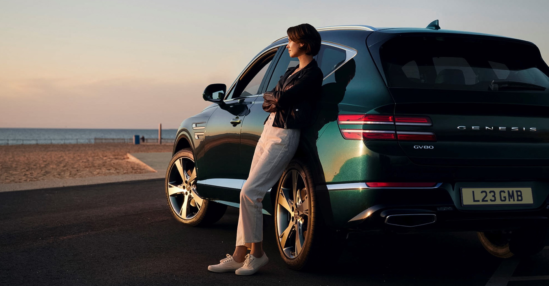 A woman contentedly leans against her Genesis SUV parked by a beach, she looks out across the sea as the sun begins to set