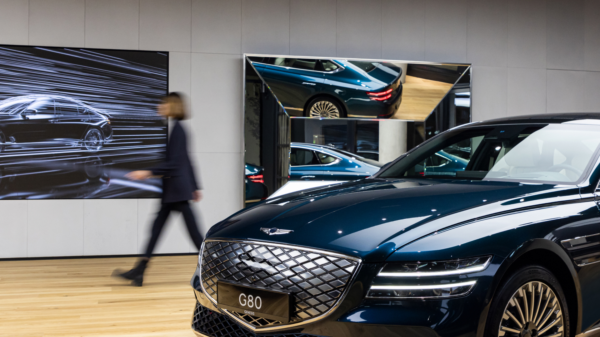 A guest walks behind Genesis G80. On the wall is a photo of a Genesis vehicle's side profile and a polyhedral mirror.