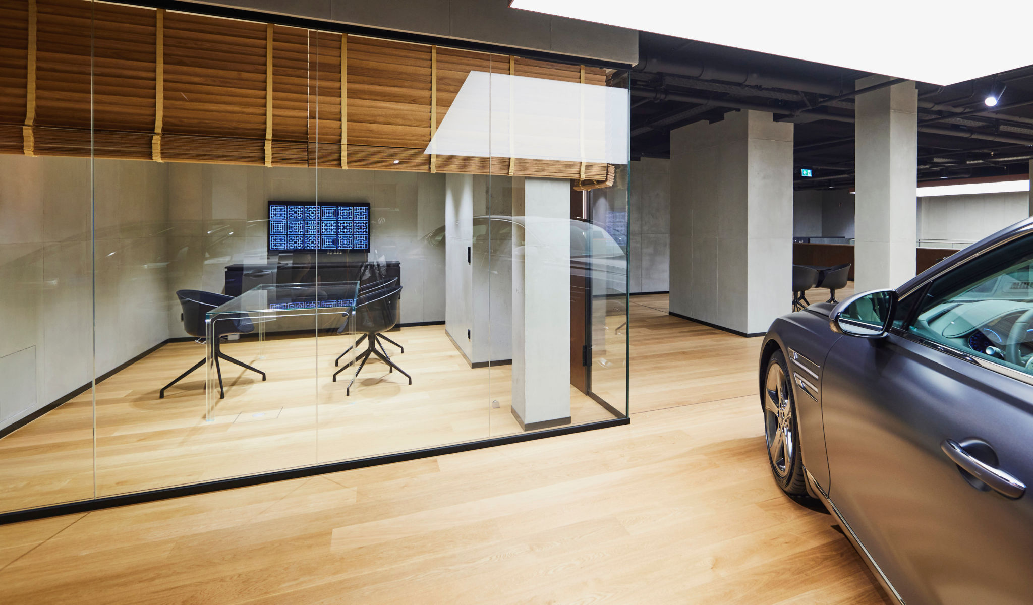 'A consultation room with glass walls and half-raised wooden blinds. Outside of the room is a partial view of a Genesis vehicle.
