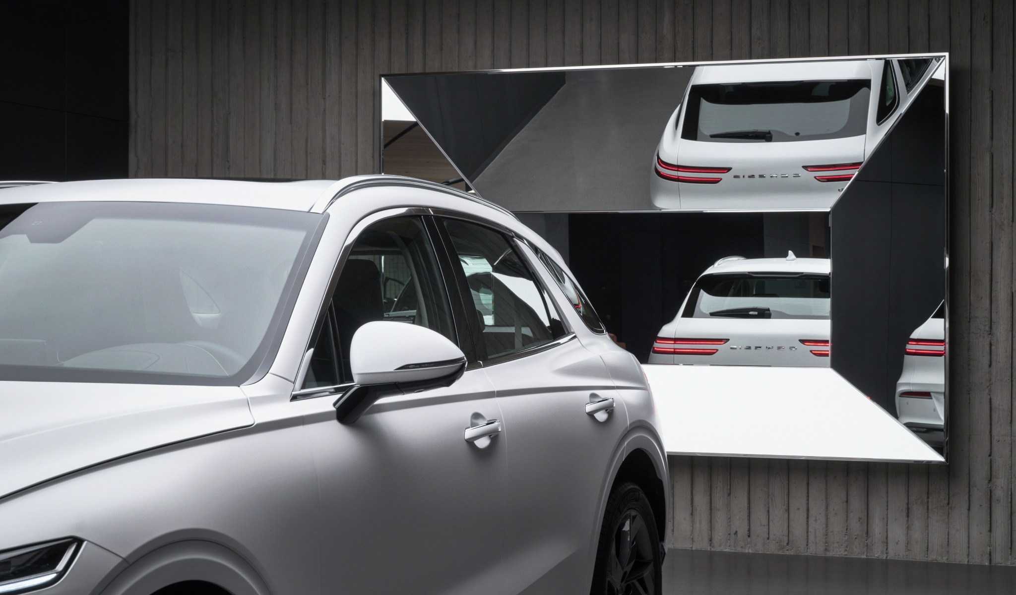 A white Genesis vehicle is on display. Behind it is a polyhedral mirror that shows the vehicle's rear in its reflection.