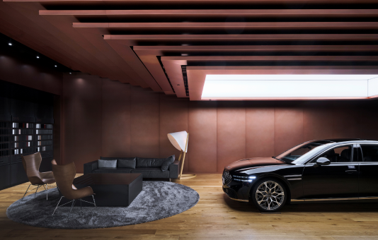 'Genesis vehicles are on display underneath lights inside a building made of weathered steel. A chair, sofa, and table lie in front of them on top of a rug.